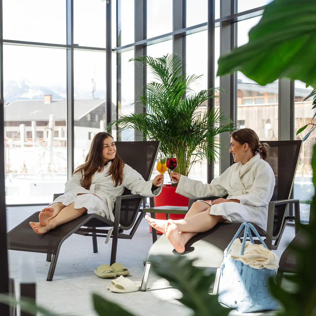 Zwei Frauen in weißen Bademänteln sitzen in Liegestühlen und halten Weingläser.